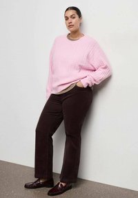 Maglione in rosa chiaro a trecce, abbinato a pantaloni in corduroy marroni. Calzature sono mocassini color bordeaux scuro. Il modello si appoggia a una parete neutra.