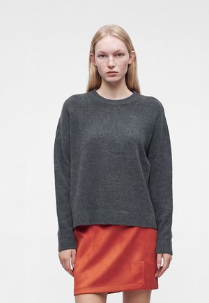 Jeune femme aux longs cheveux blonds portant un pull gris foncé et une jupe courte orange, debout devant un fond clair uni.