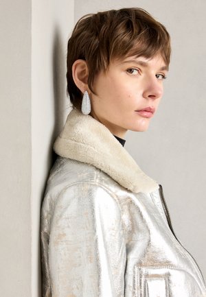 Hõbedane metallik nahktagi, millel on villane kaelus, lukuga sulgemine ja tekstuurpind. Suured tilga kujulised kõrvarõngad selgete gemmidega.