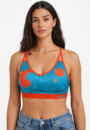Femme aux longs cheveux foncés portant un soutien-gorge de sport à motifs géométriques bleu et orange et un jean en denim bleu, debout devant un fond blanc.