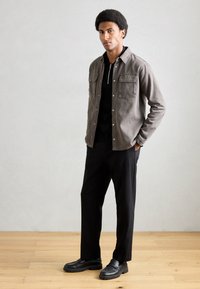 Chaqueta gris con dos bolsillos en el pecho sobre una camisa polo negra, combinada con pantalones negros y mocasines. Fondo de suelo de madera.