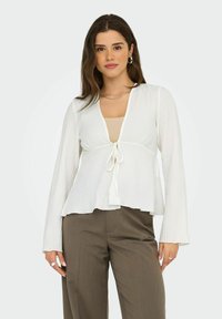Blouse blanche côtelée à manches longues, décolleté en V et détail de nœud à l'avant, associée à un top beige et un pantalon marron. Texture de tissu lisse.