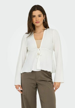 Weiße gerippte Bluse mit langen Ärmeln, V-Ausschnitt und Frontbindedetail, kombiniert mit beigem Top und braunen Hosen. Glatte Stofftextur.