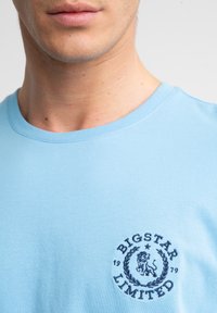 Šviesiai mėlynas medvilninis marškinėlis su apvaliu iškirpimu, pasižymintis tamsiai mėlynu siuvinėtumu, kuriame pavaizduotas liūtas ir laurų vainikas ant krūtinės.