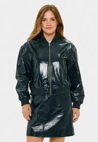 Giacca bomber lucida verde scuro con polsini e orlo a costine, dotata di tasche con zip e zip frontale. Abbinata a una gonna lucida coordinata.