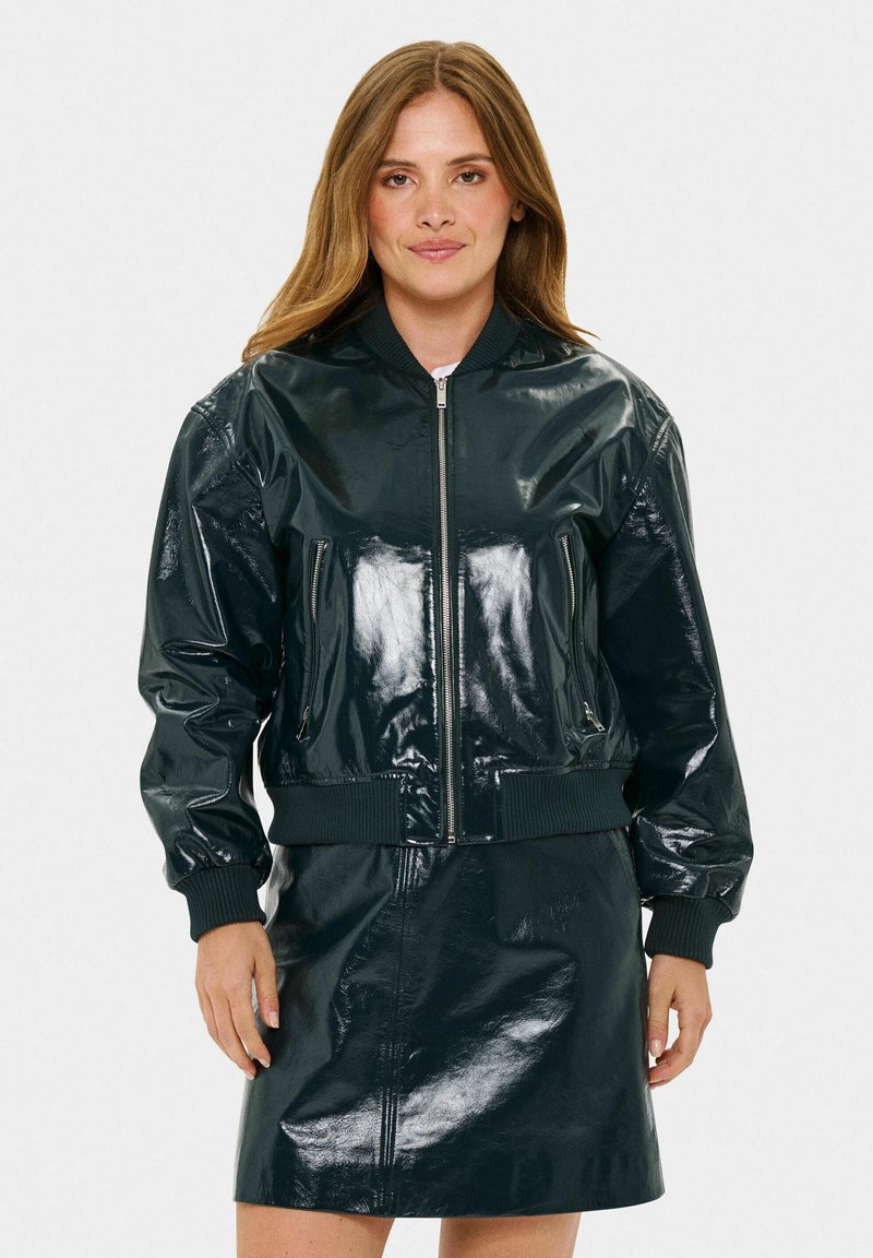 Giacca bomber lucida verde scuro con polsini e orlo a costine, dotata di tasche con zip e zip frontale. Abbinata a una gonna lucida coordinata.