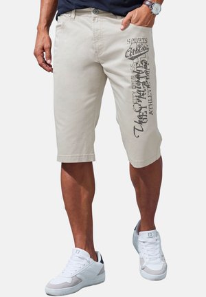 Man wearing beige knee-length shorts with black text print on right leg and white sneakers, hand in pocket, standing on white background.