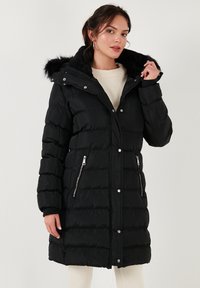 Abrigo acolchado negro con capucha de pelo, bolsillos laterales con cremallera, cierres a presión y diseño acolchado. Hecho de un material suave y resistente al agua.