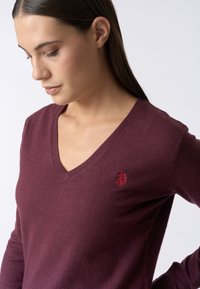 Maglione bordeaux a collo a V realizzato in morbido materiale, con un logo rosso ricamato sul lato sinistro del petto e bordi a coste sul colletto.