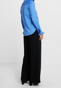 Person wearing a long-sleeve blue blouse with ruffled cuffs and wide-leg black pants, standing against a plain background.