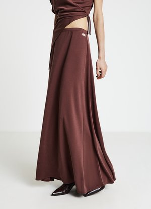 Jupe longue marron en tissu lisse et extensible. Modèle taille haute avec une fente latérale, portée avec des chaussures noires élégantes à bout pointu.