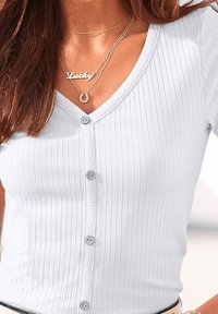 Bílý žebrovaný kardigan s výstřihem do V a pěti šedými knoflíky. Doplňují ho vrstvené zlaté náhrdelníky, včetně přívěsku "Šťastný".