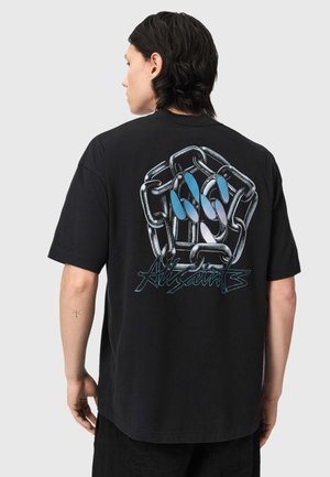 Persoană cu păr închis la culoare purtând un tricou negru cu o grafică mare de lanț și mână cu unghii albastre și textul „AllSaints” pe spate.