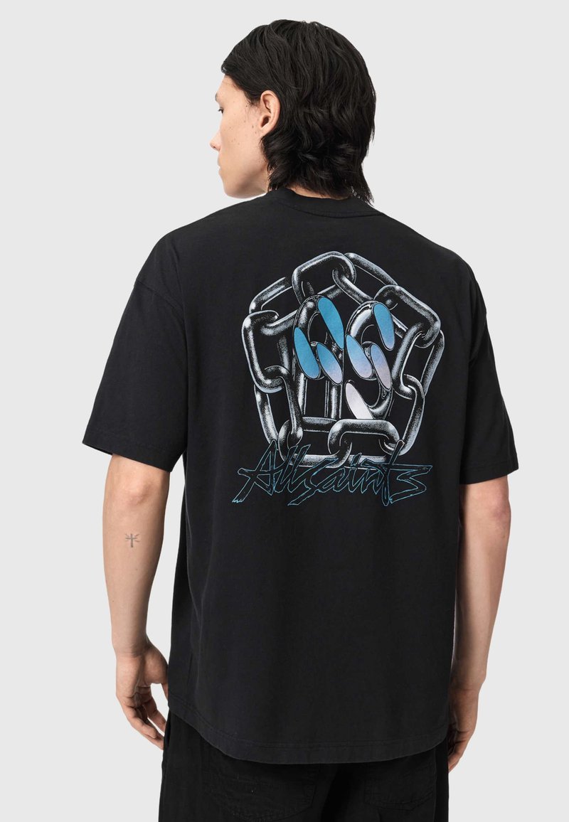 Άτομο με σκούρα μαλλιά που φορά ένα μαύρο T-shirt με μεγάλο σχέδιο αλυσίδας και χεριού με μπλε νύχια και το κείμενο «AllSaints» στην πλάτη.