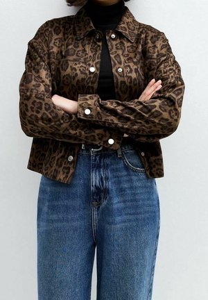 Henkilö pukeutunut ruskeaan leopardikuvioiseen takkiin mustan poolopaidan päällä, kädet ristissä ja yläosastaan näkyvissä korkea vyötäröiset siniset farkut.