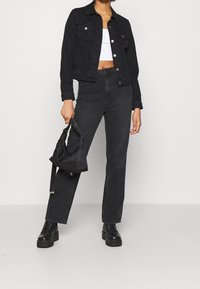 Mujer con chaqueta de mezclilla negra, pantalones de cintura alta negros, botas de plataforma negras, un top corto blanco, sosteniendo un bolso negro a su lado.