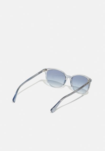Ochelari de soare tip cat-eye cu ramă transparentă de culoare albastru deschis și lentile gri cu degradé. Brațele sunt subțiri și au o finisare netedă.