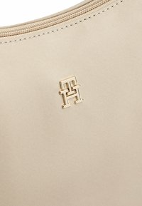 Beige stoffen tas met een gouden logo-detail, een zachte textuur en een ritssluiting langs de bovenrand.