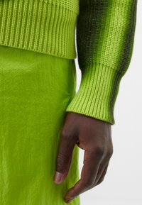 Roheline kudum, mille varrukad on soonikust, koos tekstuuriga rohelise seeliku ning heledamatest tume roheliseks üleminekuga. Seeliku ja kampsuni toonid sulanduvad harmooniliselt kokku.