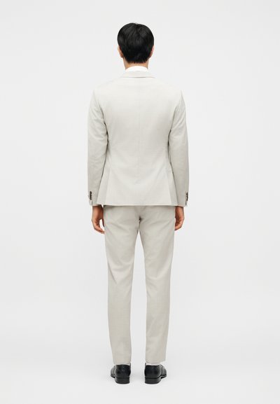 Ανοιχτό γκρι κοστούμι με ραμμένο σακάκι και αντίστοιχο παντελόνι. Λεπτή υφή, εφαρμοστό σχήμα. Μαύρα παπούτσια συμπληρώνουν το σύνολο.