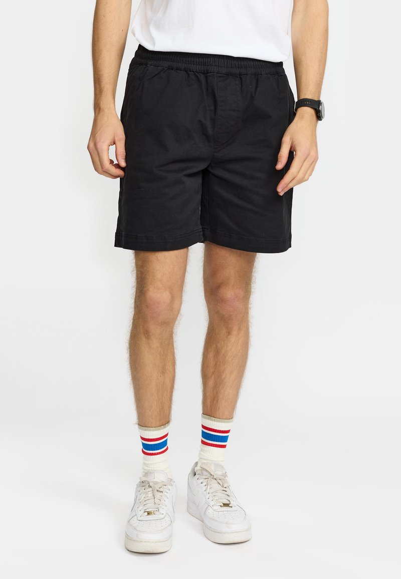 Man draagt zwarte shorts met elastische taille, witte sneakers en witte sokken met rode en blauwe strepen, staand tegen een effen witte achtergrond.