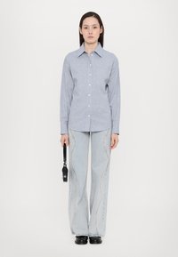 Lys blå skjorte med lange ermer, kombinert med lyse denim jeans med en bølgete dekorativ søm. Modellen holder en liten svart veske.