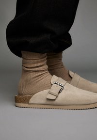 Bež semiš cipele na slip-on s detaljem kopče, korčenom potplatom i teksturiranom smeđom čarapom, uparene s labavim crnim pamučnim hlačama.