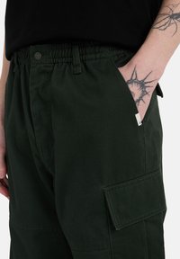 Groene cargobroek met een elastische tailleband, zijzakken en een patch pocket op de dij. Gemaakt van stevige, gestructureerde stof.