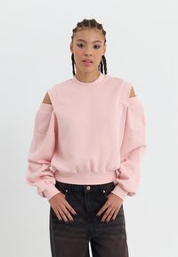 Helles rosa Sweatshirt mit Cut-Out-Schultern, Rundhalsausschnitt und Puffärmeln. Der Stoff wirkt weich; kombiniert mit dunkelblauen, hoch taillierten Jeans.