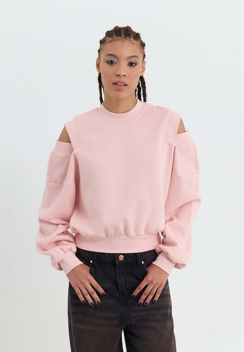 Helles rosa Sweatshirt mit Cut-Out-Schultern, Rundhalsausschnitt und Puffärmeln. Der Stoff wirkt weich; kombiniert mit dunkelblauen, hoch taillierten Jeans.