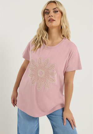 Kvinde med langt blondt hår iført en lyserød T-shirt med et stort broderet blomsterdesign og lyseblå jeans foran en ensfarvet baggrund.
