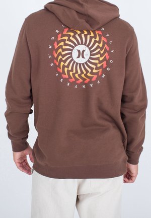 Osoba ubrana w brązową bluzę z kapturem z okrągłym wzorem promieni słońca i napisem "Hurley" na plecach, w połączeniu z jasnobeżowymi spodniami.