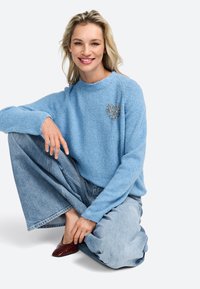 Blue fluffy sweater with a heart design in silver; paired with light blue wide-leg jeans and red pointed-toe shoes. Sitting pose.