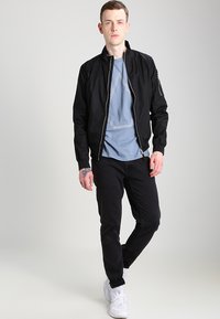Giubbotto bomber nero con una texture liscia, t-shirt grigia con un design geometrico e jeans neri. Sneakers bianche completano l'outfit.