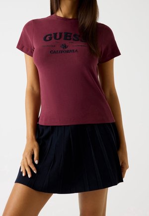 Vrouw draagt een bordeauxrode "GUESS California" T-shirt en een zwarte geplooide rok, poseert met één hand die op haar dij rust.