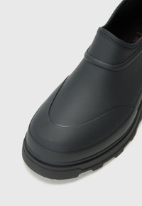 Schwarzer wasserdichter Schuh mit glattem Gummi-Obermaterial, abgerundeter Mandelform und strukturiertem Profil. Verfügt über ein niedriges Design und nahtlose Verarbeitung.