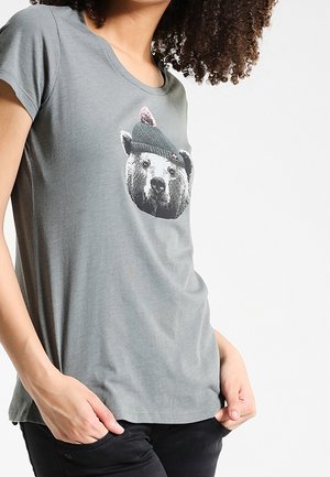Personne portant un t-shirt gris avec une illustration d'une tête d'ours portant un bonnet tricoté vert avec une plume rose.