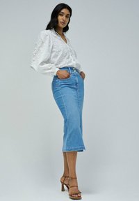 Blusa blanca con diseño floral texturizado, combinada con una falda midi de mezclilla azul claro que cuenta con una abertura frontal y dos bolsillos laterales. Sandalias de tacón marrones.