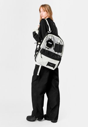 Mochila en blanco y negro con tela texturizada, múltiples bolsillos con cremallera, correas ajustables y un parche redondo en la parte frontal.