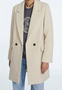 Persona vistiendo un abrigo de lana beige de doble botonadura sobre una sudadera gráfica oscura y unos vaqueros azul claro, con un collar choker negro.