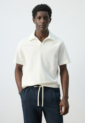 Uomo con capelli corti e ricci che indossa una polo bianca a maniche corte e pantaloni blu navy con coulisse, in piedi con le mani nelle tasche.