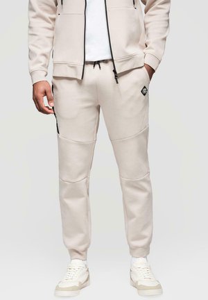 Homme portant un pantalon de jogging beige, des baskets blanches et une veste zippée beige assortie, les mains dans les poches.