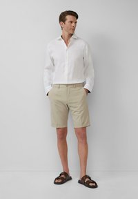 Weißes Leinenhemd, beigefarbene maßgeschneiderte Shorts und braune Sandalen. Das Hemd hat Knöpfe, die Shorts haben hochgerollte Säume und Taschen.