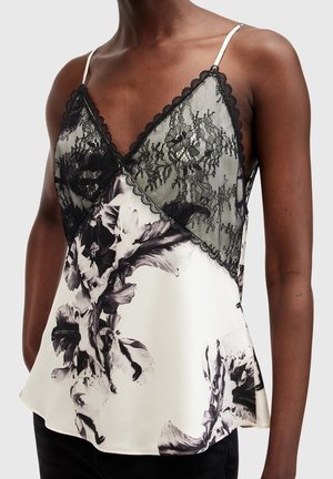 Mujer con top blanco sin mangas con estampado floral negro y paneles de encaje negro transparente con bordes ondulados en el pecho.