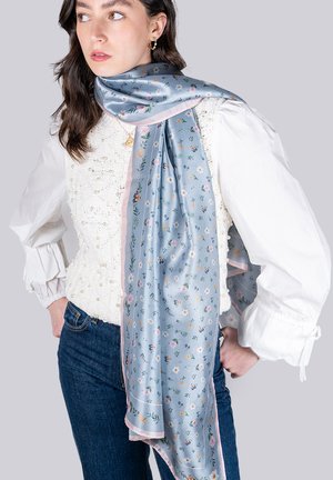 Écharpe en soie bleue avec un motif floral et des accents roses. Texture lisse, elle tombe gracieusement et complète un haut blanc texturé.