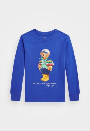Chemise bleue à manches longues avec un dessin d’un ours portant une casquette blanche, une veste rayée, un gilet vert, un pantalon bleu et des bottes jaunes.