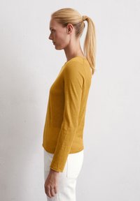 Senfgelbes Langarmoberteil mit Rundhalsausschnitt und glatter Textur, kombiniert mit weißen Hosen. Einfaches, tailliertes Design ohne Muster.