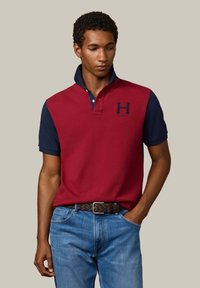 Camisa polo roja y marino con cuello clásico, mangas cortas y logo bordado de "H". Combinada con jeans azules y un cinturón texturizado marrón.