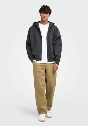 Jeune homme aux cheveux bouclés portant un sweat à capuche gris foncé zippé, un t-shirt blanc, un pantalon beige à jambes larges et des baskets blanches, debout les mains dans les poches.