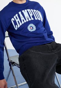 Blue sweatshirt with "CHAMPION" in white letters across the chest, round logo detail below. Worn with dark gray jeans, seated on a chair.